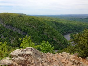 Mt. Tammany - view from summit. In the foreground is a brown rock outcropping that leads to a steep drop. Across the way there is the Delaware river meandering across the land. It is flanked by Mount Minsi. The sky is blue, mottled with clouds.