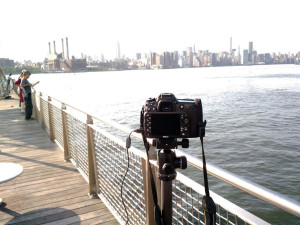 Black Nikon camera on top of a tripod. The tripod is set up on a pier in the East River of NYC. In the background there are blurred buildings along the Manhattan skyline.