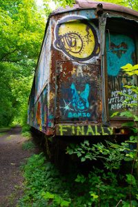 Abandoned, rusty train car in the woods covered with graffiti. There is a yellow face near the top and along the bottom is yellow-green text that says finally.