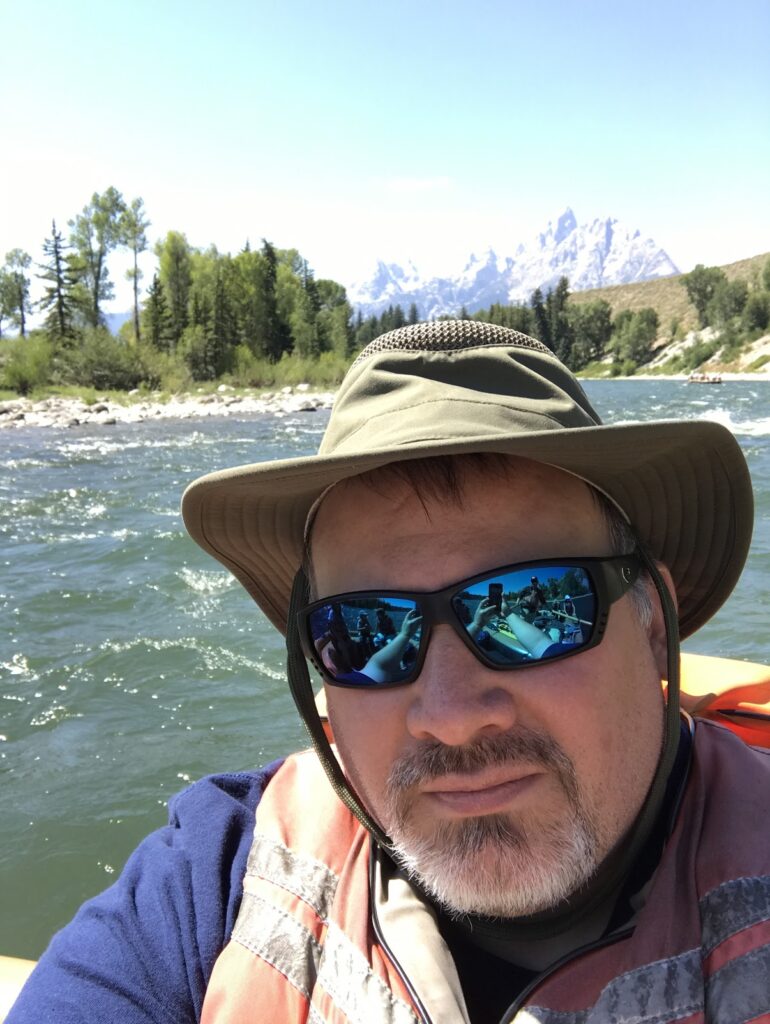 White man with salt and pepper beard and mustache wearing a brimmed olive-colored hiking hat, blue mirrored sun glasses, and an orange floatation vest. In the background is the Snake River, with some evergreen trees and the Grand Teton mountains in the background.