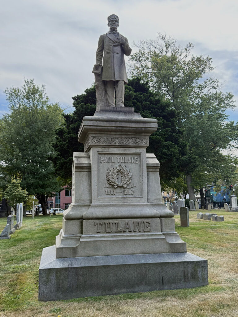 Grave marker shaped like a pedestal with a standing statue of Paul Tulane on top. The figure is wearing a buttoned up coat and a hat. His right had is seated on top of a tree stump.